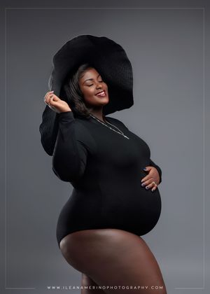 Smiling pregnant woman in black bodysuit and wide-brim hat cradles baby bump in a professional maternity portrait.