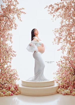 Pregnant woman in white off-shoulder gown stands on pedestal framed by pink cherry blossoms.