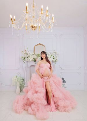 Smiling pregnant woman in pink tulle gown stands under crystal chandelier in white studio.