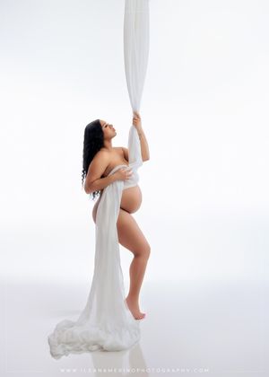 Pregnant woman draped in white fabric poses gracefully against a bright white background.