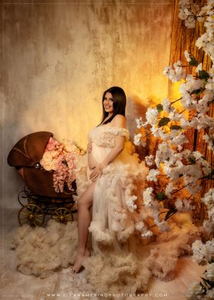 Pregnant woman in cream tulle gown poses beside vintage pram and blooming floral trees.