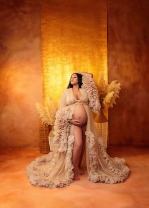 Glamorous maternity photo of pregnant woman in ruffled champagne gown against golden backdrop.
