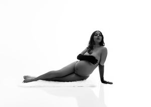 Black and white maternity photo of pregnant woman posing gracefully on white fur rug in studio.