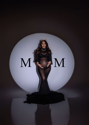 Dramatic maternity portrait of pregnant woman in black gown with crown before MOM spotlight.