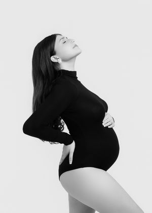 Black and white maternity portrait of pregnant woman in black bodysuit against white background.