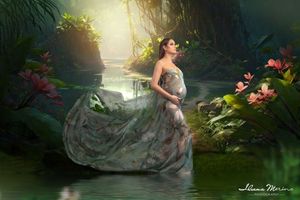 Pregnant woman in flowing floral gown standing in enchanted forest with water and tropical flowers.