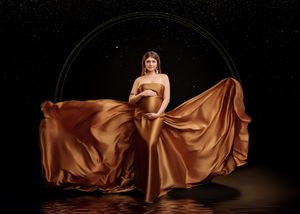 Pregnant woman in elegant gold satin gown with circular hoop backdrop on reflective dark surface.