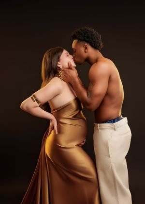 Pregnant woman in gold satin gown shares intimate moment with shirtless partner in studio.