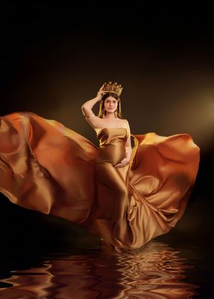Pregnant woman wearing gold crown and flowing copper satin gown reflected in water on dark background.