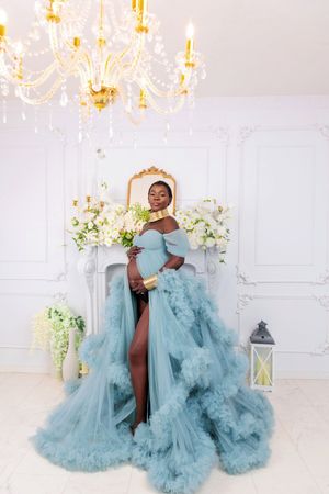 Pregnant Black woman in ruffled teal tulle gown posing near floral fireplace with chandelier.