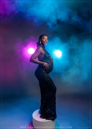 Pregnant Black woman in black sequin gown posing on pedestal with colorful smoke studio backdrop.