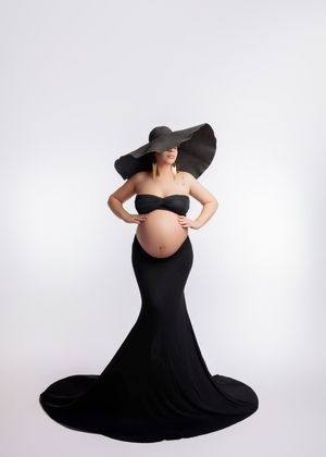 Pregnant woman in strapless black mermaid gown wearing oversized black sun hat on white background.