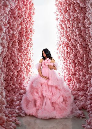 Pregnant woman in an elegant pink ruffled gown stands between walls of pink roses in a stunning maternity photo.