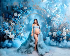 Pregnant woman in a sheer blue gown poses before a stunning floral arch with blue flowers and dreamy cloud-like backdrop.