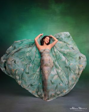 Pregnant woman draped in a flowing green floral fabric gown poses gracefully against a green studio backdrop.