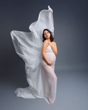 Pregnant woman draped in flowing white sheer fabric poses gracefully against a gray background in a maternity portrait.
