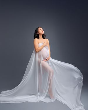 Pregnant woman wrapped in flowing white sheer fabric poses elegantly against a gray studio background in maternity photo.