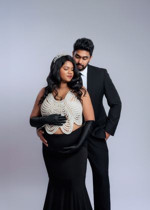 Pregnant woman in pearl bodice and crown embraced by partner in black suit for maternity photo.