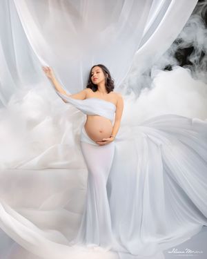 Pregnant woman in white gown poses with billowing fabric and smoke in an ethereal maternity photography studio setting.
