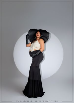 Pregnant woman in black skirt and wide-brim hat poses elegantly against a white circular backdrop in maternity photo.