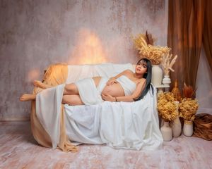 Pregnant woman in white lingerie reclines on a draped sofa surrounded by dried flowers in a warm maternity photo.