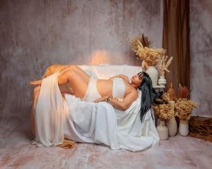 Pregnant woman in white lingerie reclines on draped sofa surrounded by dried floral arrangements.