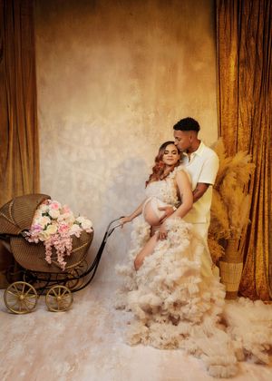 Couple poses with vintage floral pram, woman in ruffled maternity gown against golden backdrop.