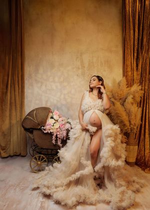 Pregnant woman in ruffled champagne gown sits beside vintage floral pram in golden studio setting.