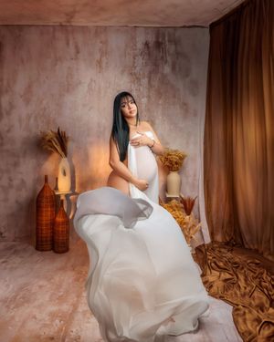 Pregnant woman in flowing white fabric poses in warmly lit studio with decorative vases.
