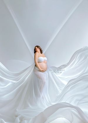 Pregnant woman in white strapless gown surrounded by dramatic flowing white fabric in studio.