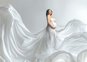Pregnant woman in flowing white silk gown with billowing fabric against soft grey background.