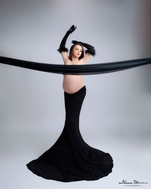 Pregnant woman in black mermaid skirt and gloves poses with flowing black fabric in studio.