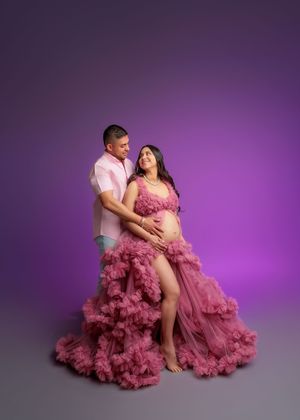 Couple in pink outfits share tender moment in romantic purple-backdrop maternity session.