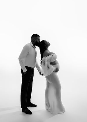 Black and white maternity portrait of couple sharing kiss with woman in elegant white gown.