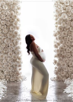 Pregnant woman in white dress posed between two tall white rose floral columns in bright studio.