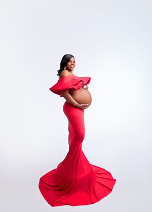 Pregnant woman in stunning red off-shoulder gown with long train against white studio background.