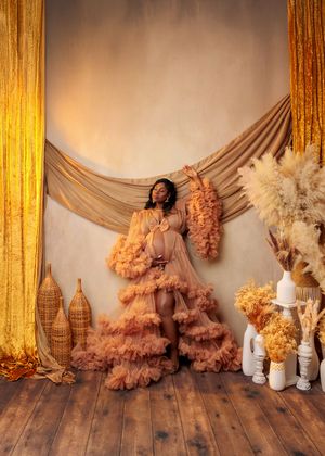 Pregnant woman in peach ruffled maternity gown posing in boho-styled studio with warm golden tones.