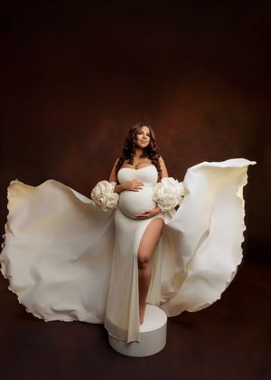 Pregnant woman in white flowing gown with large rose accents and billowing fabric in studio.