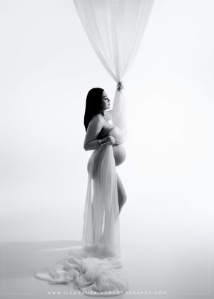 Black and white artistic maternity photo of pregnant woman holding sheer white fabric drape.