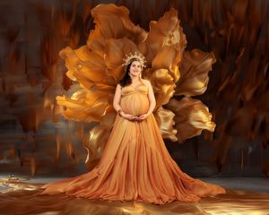 Pregnant woman in flowing golden orange gown with crown poses against dramatic golden floral backdrop.