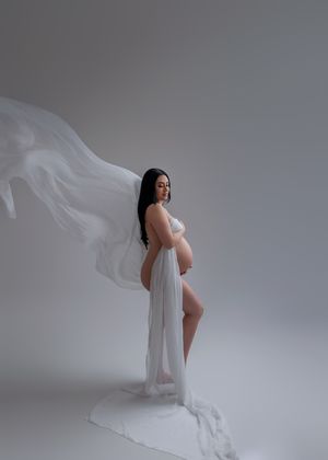 Pregnant woman draped in flowing white sheer fabric poses gracefully against a soft grey studio background.