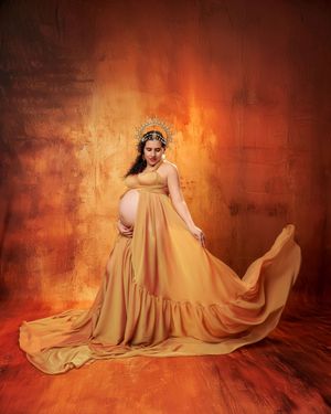 Pregnant woman wearing golden crown and mustard yellow gown with train poses on warm textured background.