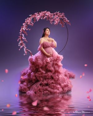 Pregnant woman in pink ruffled gown sits on floral hoop swing with cherry blossoms falling around her.