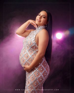 Pregnant woman in sparkling crystal embellished sheer gown poses against dark smoky purple lit background.