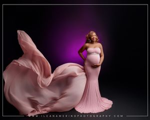 Pregnant woman in pink strapless mermaid gown with flowing fabric train poses against dark purple backdrop.
