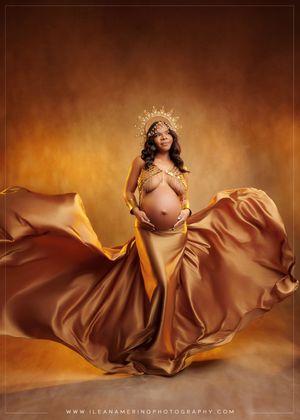 Pregnant woman in gold satin flowing gown with crown poses in elegant maternity portrait.