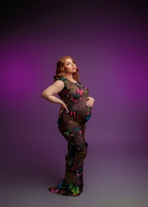 Pregnant woman in colorful butterfly floral sheer gown poses against purple backdrop in maternity photo.