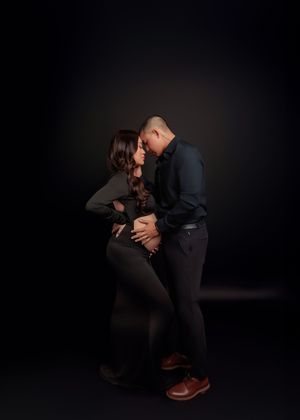 Couple sharing intimate maternity moment, hands on baby bump, dark studio background.
