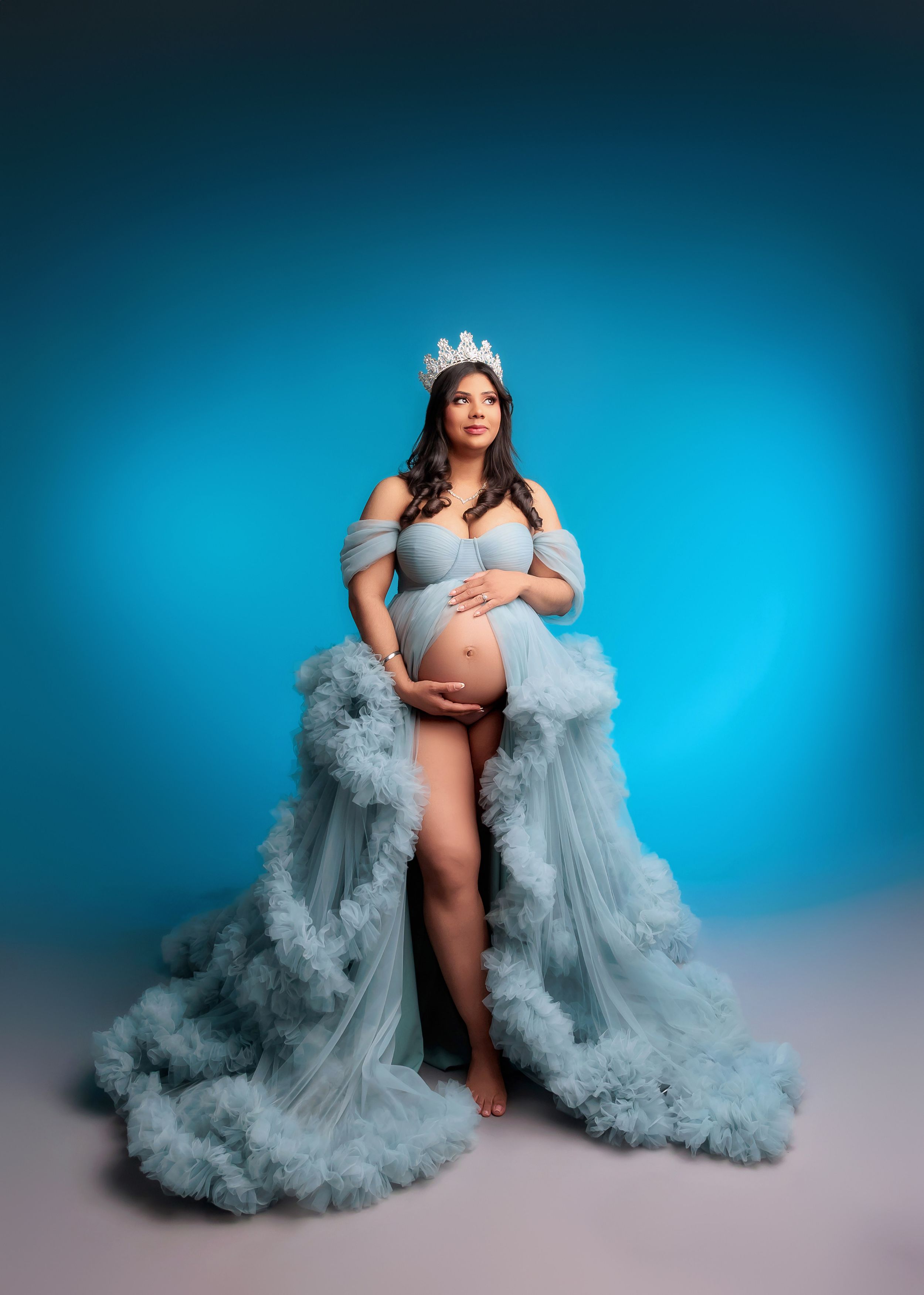 Pregnant woman wearing crown in blue ruffled tulle gown against blue gradient backdrop.