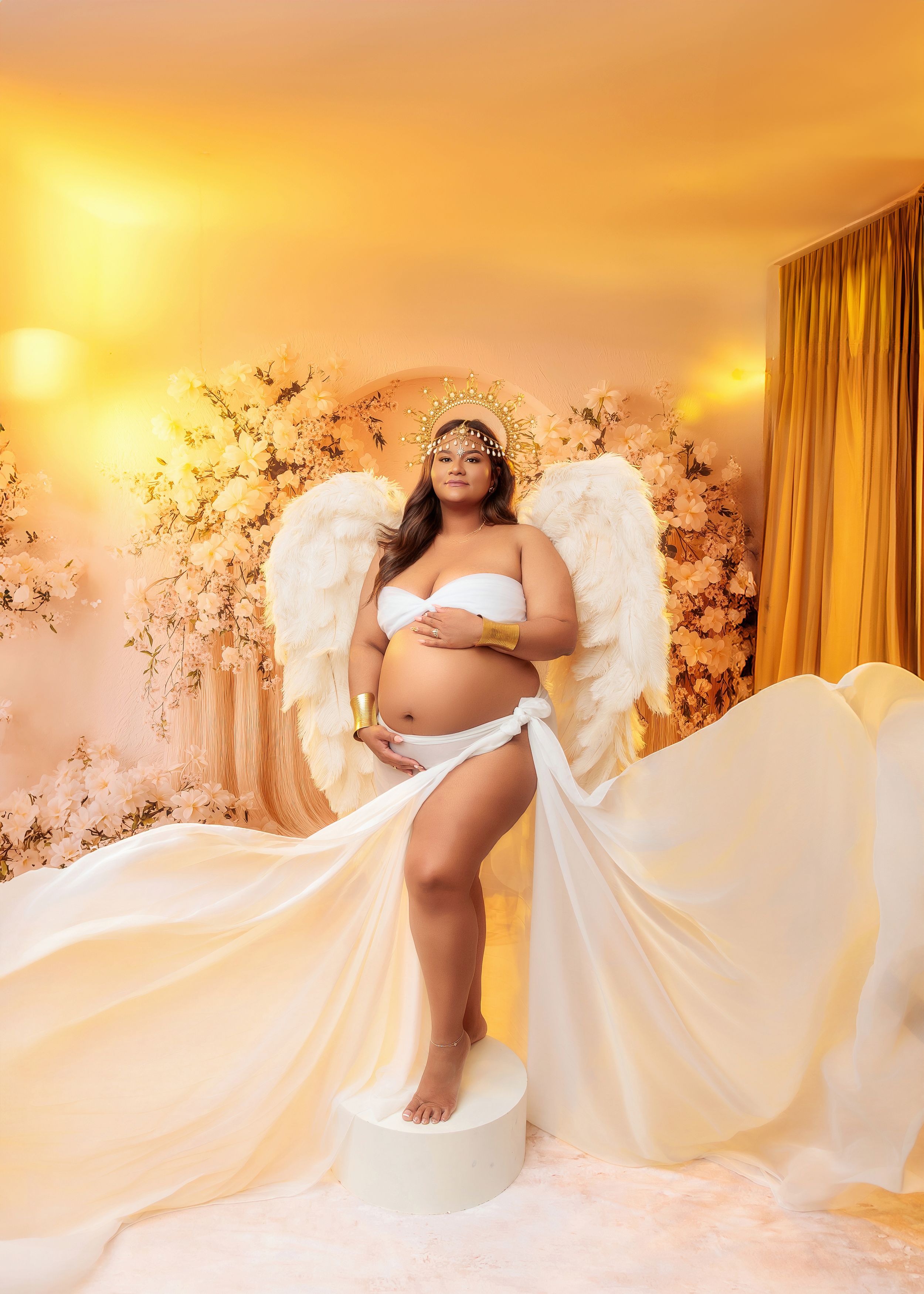 Pregnant woman in angel wings and white flowing fabric, golden floral backdrop.